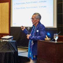 Dr. Komatsu speaking into a microphone at a podium with powerpoint slides on a projector