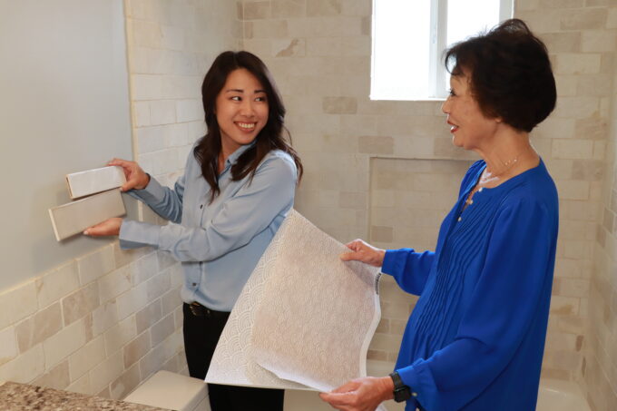 Desiree Kitagawa holding up tile samples and Beverly Ito hold wallpaper samples looking at each other
