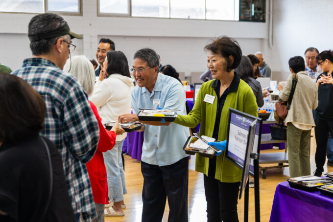 a man in blue and a woman in green handing out bentos