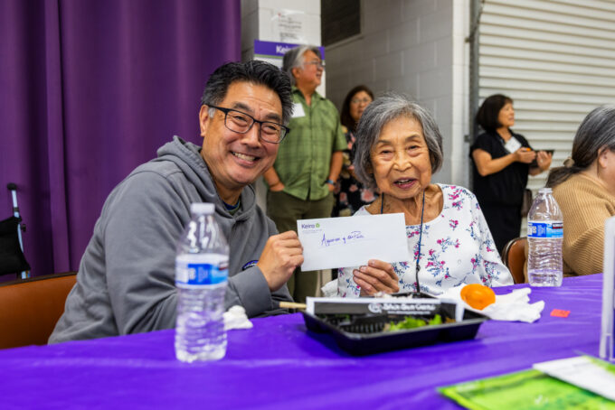 a man in gray and a woman in a white floral shirt holding up an envelope