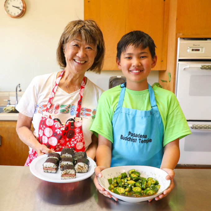 grandma and grandson holding plates of spam musubi and cucumber salad