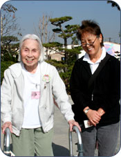 An adult woman stands next to an adult senior woman with a walker.