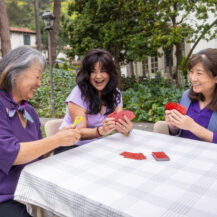 three women wearing purple playing cards outside at a table