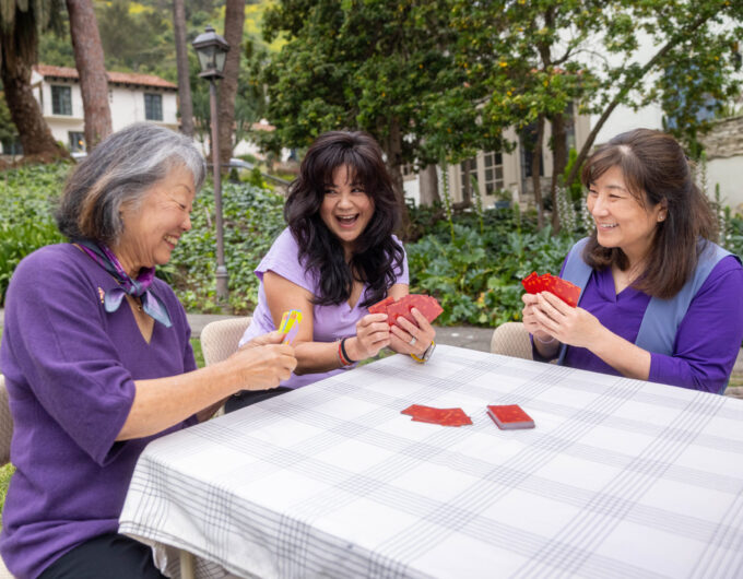 three women wearing purple playing cards outside at a table