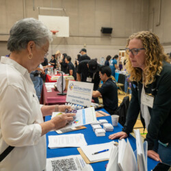 a woman in a black jacket blonde curly hair talking with an older lady in a white button up