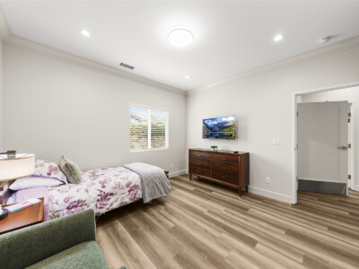a bed with purple and white bedding, a chair, wood dresser, and tv in a bedroom