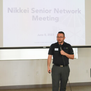 man talking into microphone, presentation behind him reads "Nikkei Senior Network Meeting"