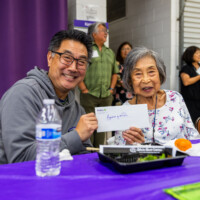 a man in gray and a woman in a white floral shirt holding up an envelope